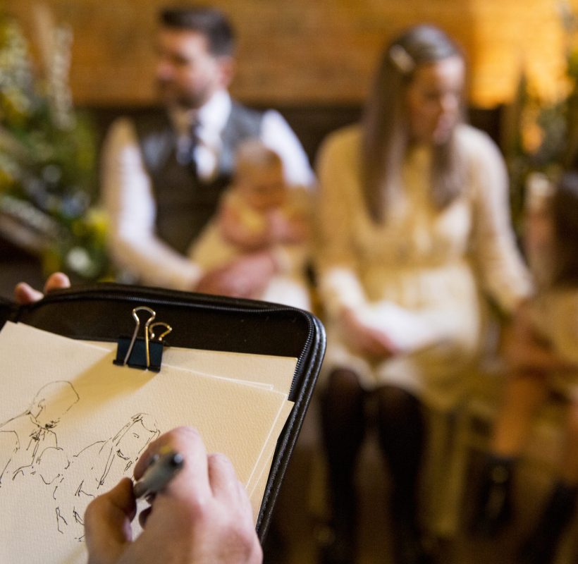 Over the shoulder view of artist sketching family during naming ceremony in an historic barn.