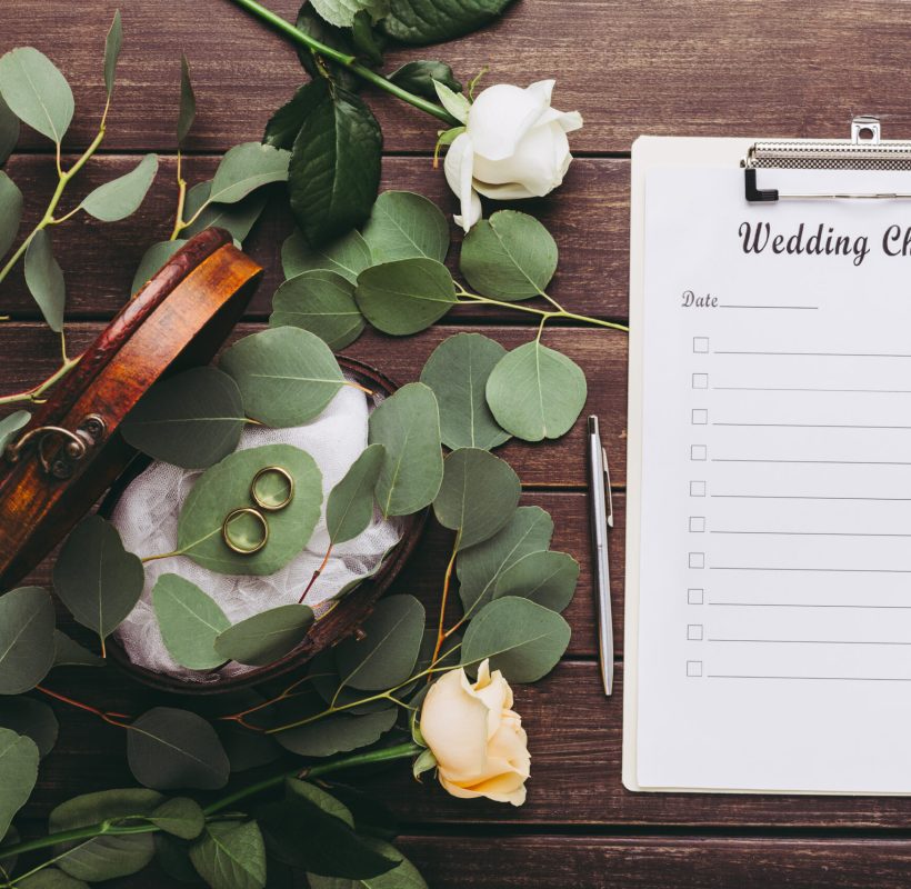 Marriage concept. Wedding rings and paper planning checklist on wooden table with green natural leaves, copy space